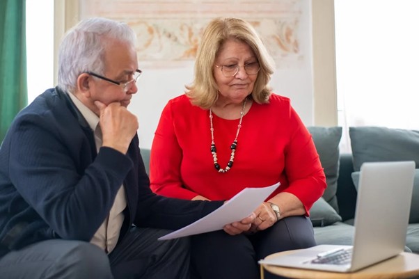 A man and a woman sitting on a sofa while looking at paper and a laptop.