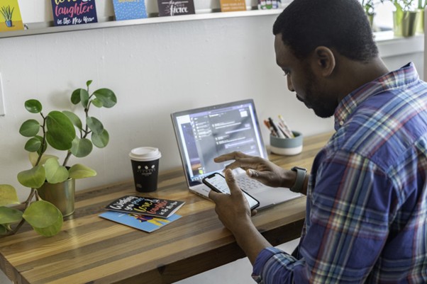 Man sitting at a desk with a laptop while looking at a mobile phone.