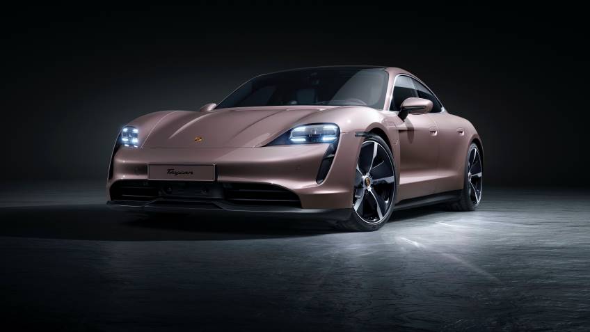 Rose gold 4 door Porsche Taycan with lights on parked under a spotlight with everything else blacked out